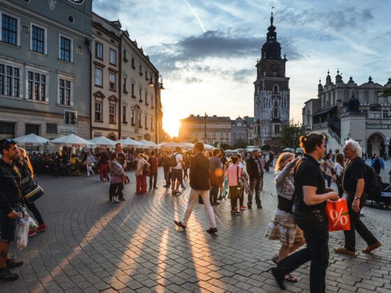 Crowded European city square at sunset with cafes and historic buildings, a tall clock tower in the background under a blue sky.
