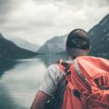 Person wearing an orange hiking backpack and striped shirt seen from behind, overlooking a calm lake with mountains in the distance under cloudy skies. It appears to be a outdoor adventure or hiking scene.