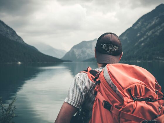 Person wearing an orange hiking backpack and striped shirt seen from behind, overlooking a calm lake with mountains in the distance under cloudy skies. It appears to be a outdoor adventure or hiking scene.