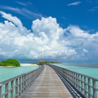 Wooden boardwalk extends over turquoise water toward thatched huts on a distant island under a blue sky with white clouds.