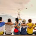 Group of friends sitting shoulder-to-shoulder on a railing by the water, arms around each other, watching the sea with cable cars and mountains in the background at sunset.
