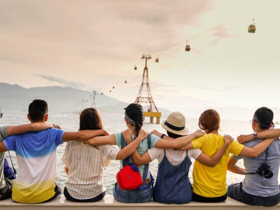 Group of friends sitting shoulder-to-shoulder on a railing by the water, arms around each other, watching the sea with cable cars and mountains in the background at sunset.