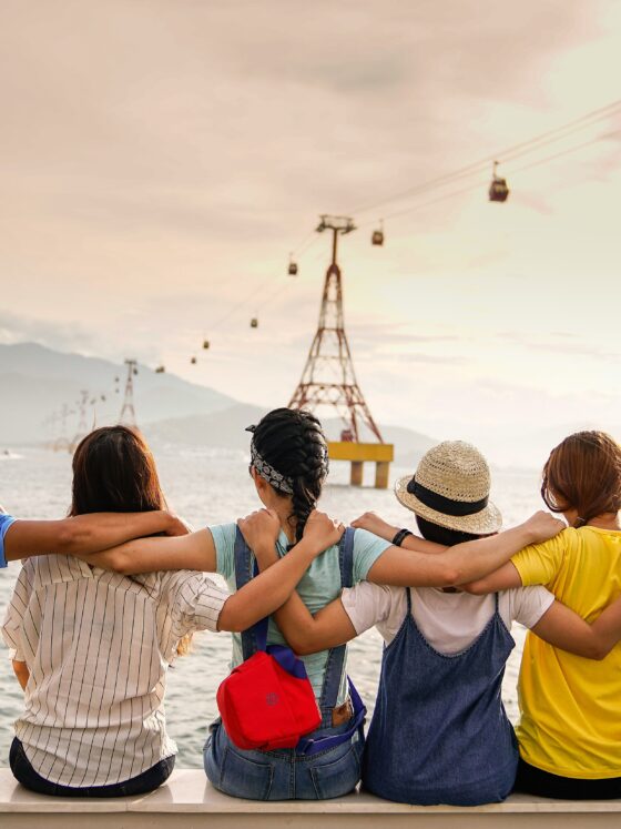 Group of friends sitting shoulder-to-shoulder on a railing by the water, arms around each other, watching the sea with cable cars and mountains in the background at sunset.