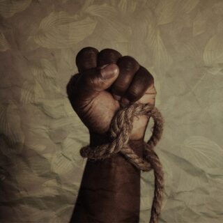 A dark-skinned hand clenched into a fist, bound with rough rope around the wrist, against a leaf-patterned background.