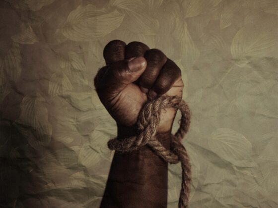 A dark-skinned hand clenched into a fist, bound with rough rope around the wrist, against a leaf-patterned background.