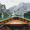 View from the bow of a wooden boat on a calm alpine lake, with steep rocky mountains and pine-covered shores in the background.