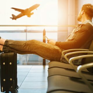 Man relaxing in an airport terminal with feet on a suitcase while a plane takes off outside the window.