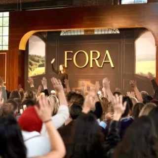 Speaker on a stage at a conference with a large FORA sign in the background, crowd raising hands in an indoor venue.