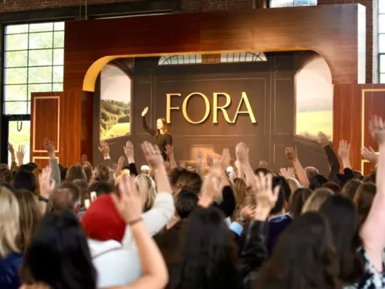 Speaker on a stage at a conference with a large FORA sign in the background, crowd raising hands in an indoor venue.