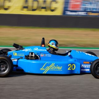 Blue Formula-style race car speeding on the track, driver in helmet, car number 20 visible and sponsor decals along the side.