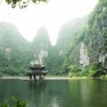 Traditional Chinese pavilion on a calm lake, framed by towering green cliffs and misty mountains.