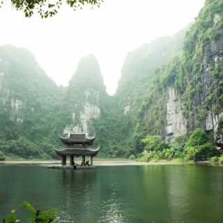Traditional Chinese pavilion on a calm lake, framed by towering green cliffs and misty mountains.