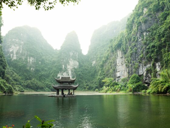 Traditional Chinese pavilion on a calm lake, framed by towering green cliffs and misty mountains.