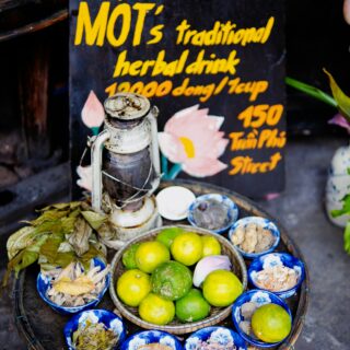 Basket of limes and blue bowls with dried ingredients on a market table, with a dark sign featuring yellow Vietnamese text in the background.