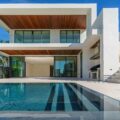 Modern white, two-story house with glass balcony and a blue reflecting pool in front under a clear sky.