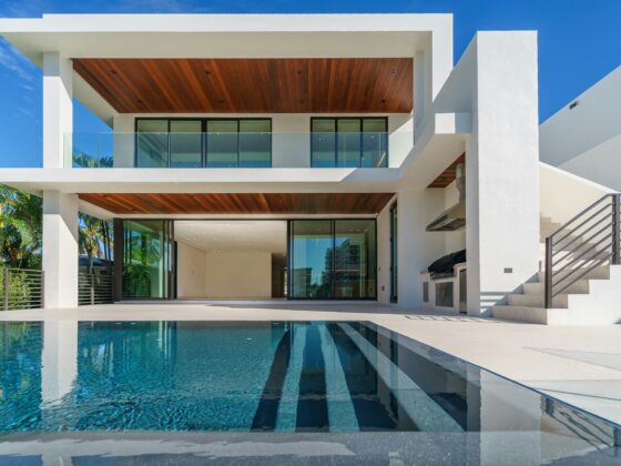 Modern white, two-story house with glass balcony and a blue reflecting pool in front under a clear sky.
