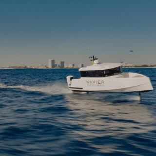 White electric flying boat cutting through blue water with a city skyline in the distance; 'NAVIER' logo visible on hull.