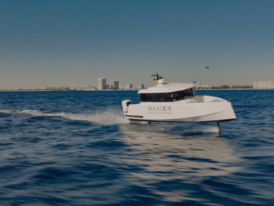 White electric flying boat cutting through blue water with a city skyline in the distance; 'NAVIER' logo visible on hull.