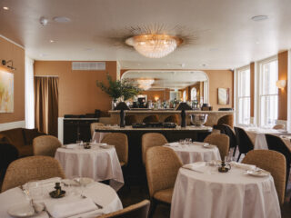 Elegant dining room with round tables in white linens, wicker chairs, and a crystal chandelier overhead at the bar area