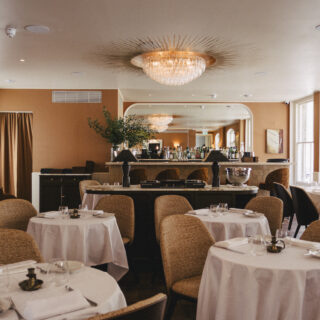 Elegant dining room with round tables in white linens, wicker chairs, and a crystal chandelier overhead at the bar area