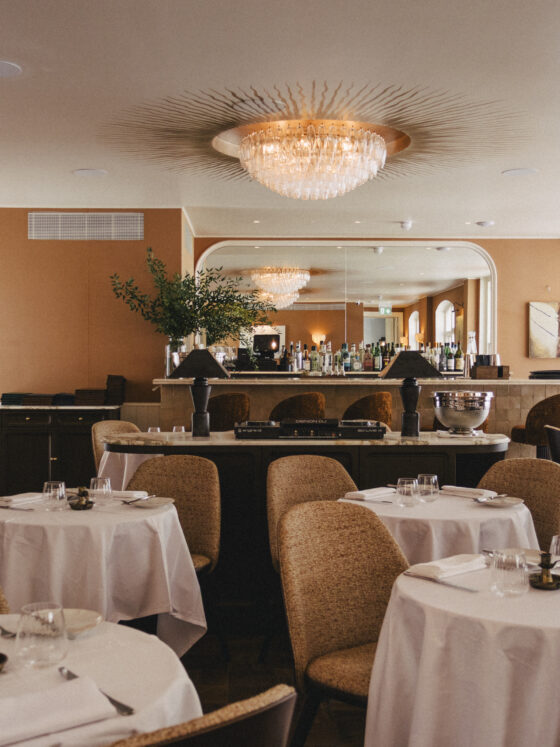 Elegant dining room with round tables in white linens, wicker chairs, and a crystal chandelier overhead at the bar area