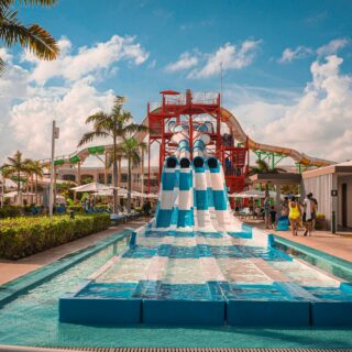 Water park with a tall red platform and blue-and-white slides, surrounded by sunlit pool and palm trees, with people nearby.
