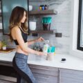 Athletic woman in a sports bra scooping powder into a cup on a kitchen counter; protein shake packets on a shelf behind her.