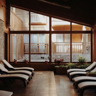 Sunlit indoor spa lounge with curved wooden ceiling, stone wall, and rows of white lounge chairs along large glass windows.