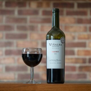 Wine bottle labeled Viñailba Malbec and a filled red wine glass on a wooden table with a brick wall in the background.