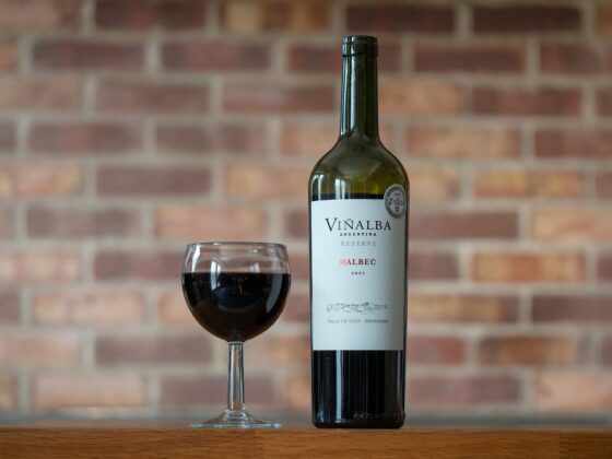 Wine bottle labeled Viñailba Malbec and a filled red wine glass on a wooden table with a brick wall in the background.