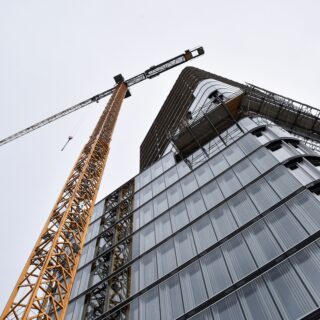 Construction crane towers beside a modern glass building under construction, steel framework visible against a pale sky