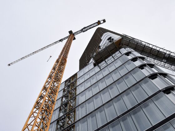 Construction crane towers beside a modern glass building under construction, steel framework visible against a pale sky