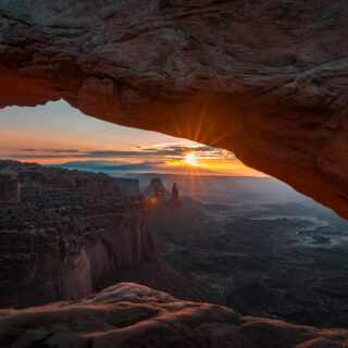 Sunset through a natural rock arch over a rugged canyon, warm orange light illuminating the cliffs.