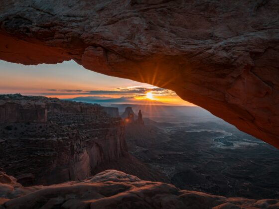 Sunset through a natural rock arch over a rugged canyon, warm orange light illuminating the cliffs.