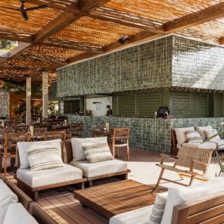 Open-air lounge with beige-cushioned wooden sofas and chairs around a low coffee table, beside a green-tiled bar under a woven roof.