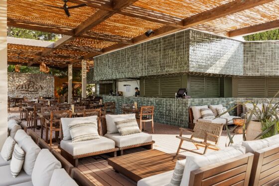 Open-air lounge with beige-cushioned wooden sofas and chairs around a low coffee table, beside a green-tiled bar under a woven roof.