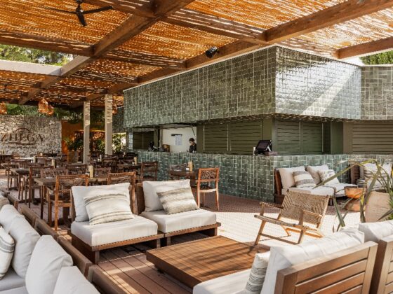 Open-air lounge with beige-cushioned wooden sofas and chairs around a low coffee table, beside a green-tiled bar under a woven roof.
