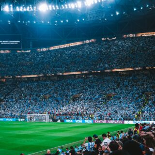 A packed stadium with a green football pitch, banners reading FIFA World Cup Qatar 2022, and a sea of cheering fans in blue and white.