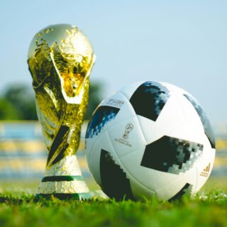 Soccer ball beside a gold trophy on a grassy field under a clear sky.