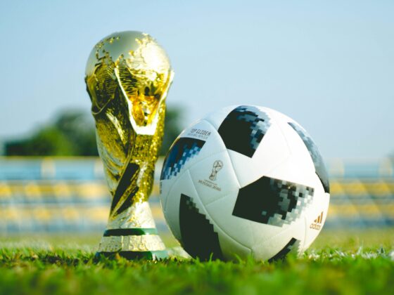 Soccer ball beside a gold trophy on a grassy field under a clear sky.