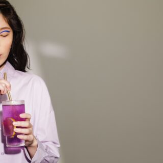 Woman with blue eyeliner wearing a lavender shirt sipping a purple drink through a straw with eyes closed.