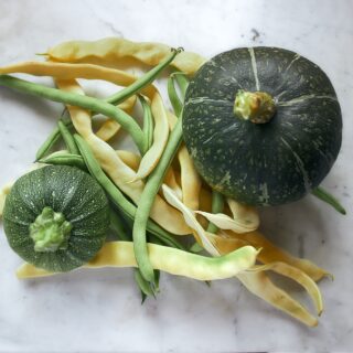 Two dark green pumpkins with pale streaks lie among green and yellow beans on a white marble surface.