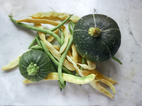 Two dark green pumpkins with pale streaks lie among green and yellow beans on a white marble surface.