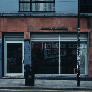 Storefront with a large glass window and door, orange neon sign reading 'CUBINTS' above the entrance, a black trash bin on the sidewalk, and a sticker-covered street pole in front.