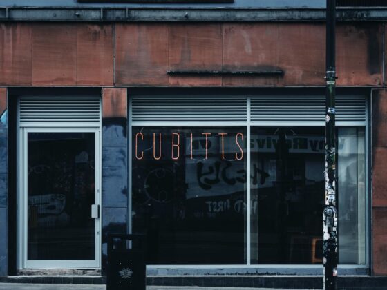 Storefront with a large glass window and door, orange neon sign reading 'CUBINTS' above the entrance, a black trash bin on the sidewalk, and a sticker-covered street pole in front.