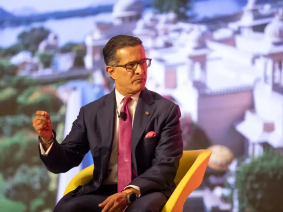 Man in a navy suit and pink tie seated in a yellow chair on stage, speaking with a lapel microphone.
