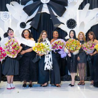 Group of ten women in evening gowns holding colorful bouquets on a stage with a white decorative backdrop of flowers and wings.