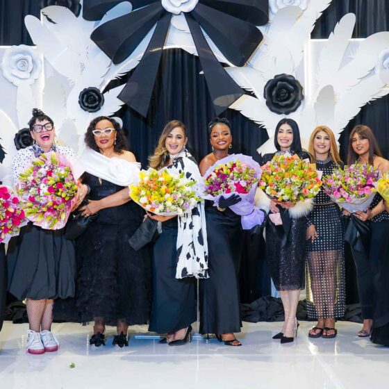 Group of ten women in evening gowns holding colorful bouquets on a stage with a white decorative backdrop of flowers and wings.