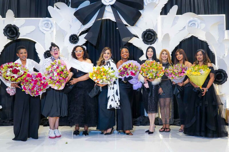 Group of ten women in evening gowns holding colorful bouquets on a stage with a white decorative backdrop of flowers and wings.