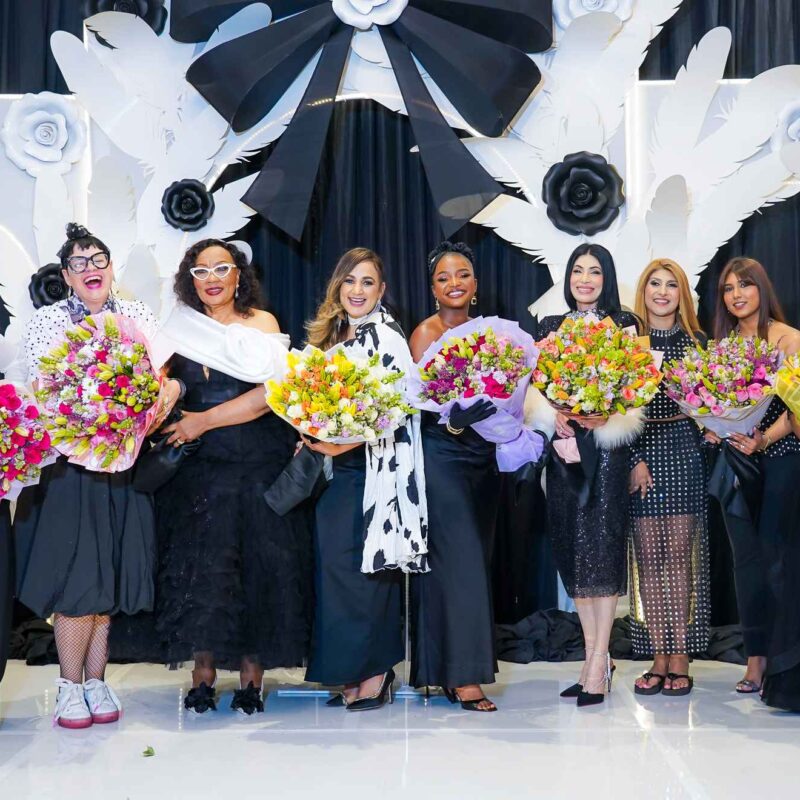 Group of ten women in evening gowns holding colorful bouquets on a stage with a white decorative backdrop of flowers and wings.